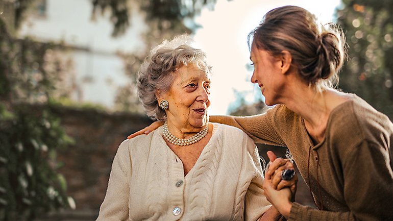 En medelålders kvinna håller om en äldre kvinna och de kollar på varandra och ler.