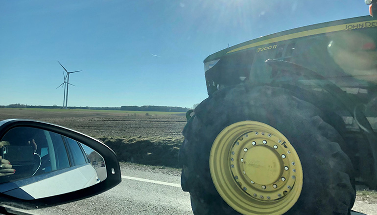 Traktor fotograferad från en bil. Vindkraftverk syns i bakgrunden.