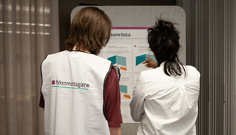 Två personer i vit väst står med ryggen mot. På västen står det röstmottagare.