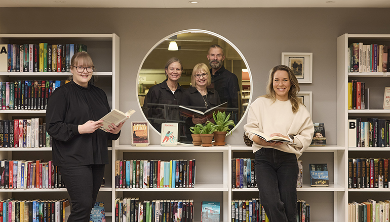 Fem personer står i ett bibliotek. Tre av personerna håller i en bok. Alla ler och tittar in i kameran.