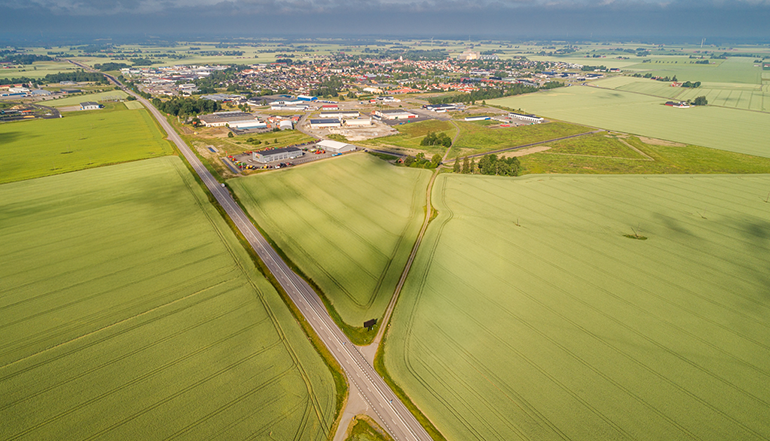 Drönarbild över åkermark med en stor väg som går igenom på mitten. I bakgrunden ett samhälle.