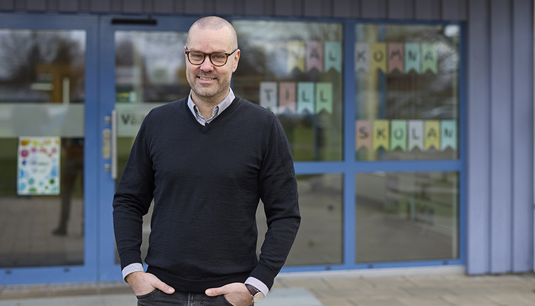 André Essebro står framför en skolentré i Vara. Han ler mot kameran.