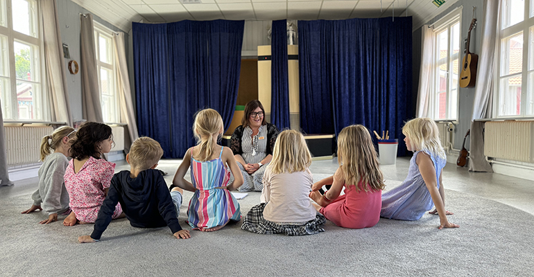 barngrupp sitter framför på golvet med ryggen vänd mot kameran, framför sitter en pedagog som ler mot barnen. I bakgrunden syns en scen i en lokal.