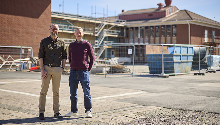 Två män stor fram en tegelbyggnad i två våningar med byggställningar framför fasaden.
