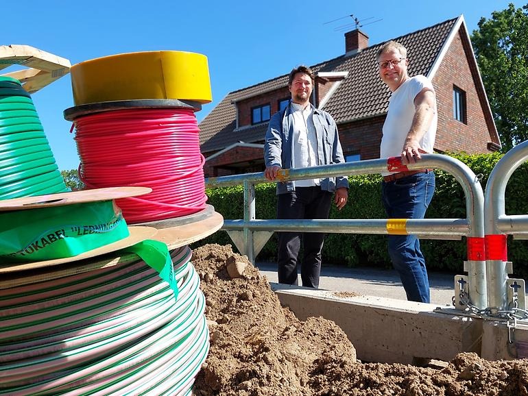 Tre kabelvindor med röda och gröna kablar i en sandhög. Bredvid finns ett staket som två män står vid.