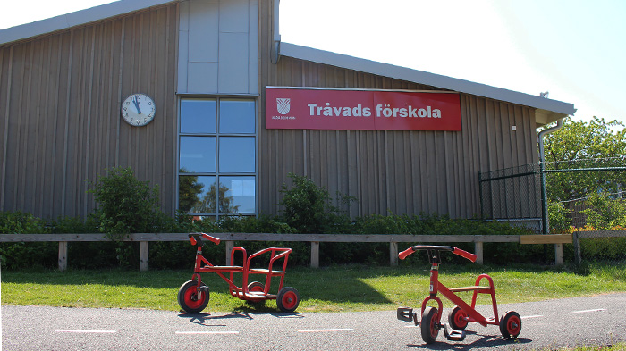 Två röda trehjulingar i förgrunden, skolbyggnad i trä i bakgrunden.