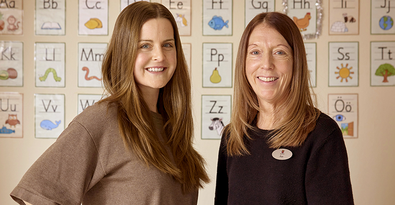 Två kvinnor står bredvid varandra. I bakgrunden syns alfabetet