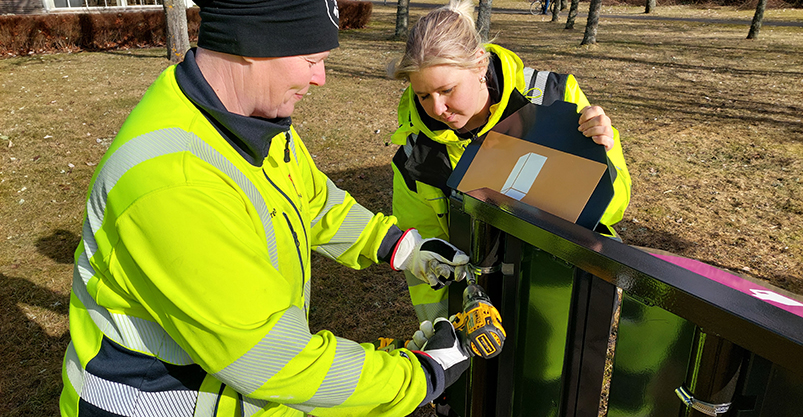 Två kvinnor i varselkläder monterar upp en behållare på en ställning.