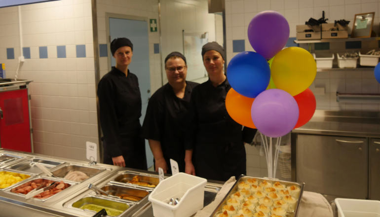 Tre personer i bakom disken i ett skolmatsalskök, ballonger på höger sida.