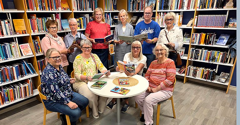 Tio personer står och sitter vid ett bord som står mitt i ett bibliotek.